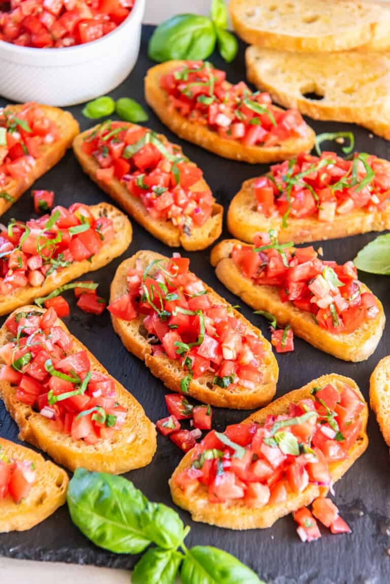 Crispy crostini topped with chopped tomatoes, garlic, onion and fresh basil, arranged on a slate serving board with basil leaves and a bowl of bruschetta mixture.