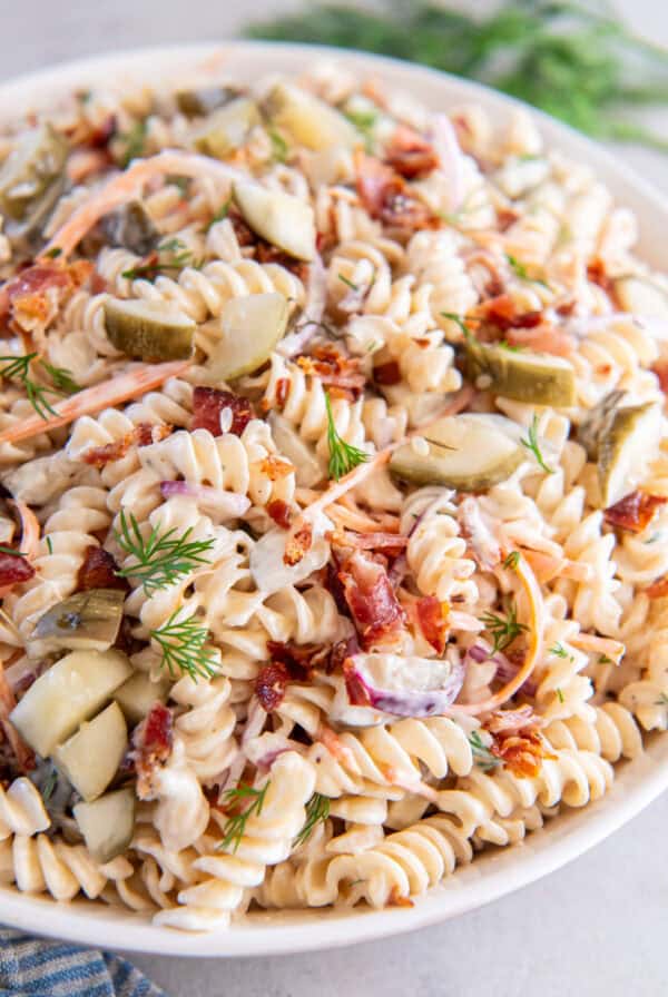 A white bowl with pasta, vegetables, and pickles covered in a creamy sauce.