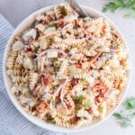 A white bowl with pasta, vegetables, and pickles covered in a creamy sauce.