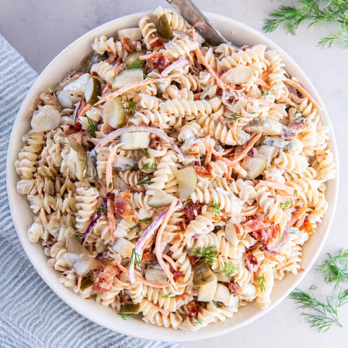 A white bowl with pasta, vegetables, and pickles covered in a creamy sauce.