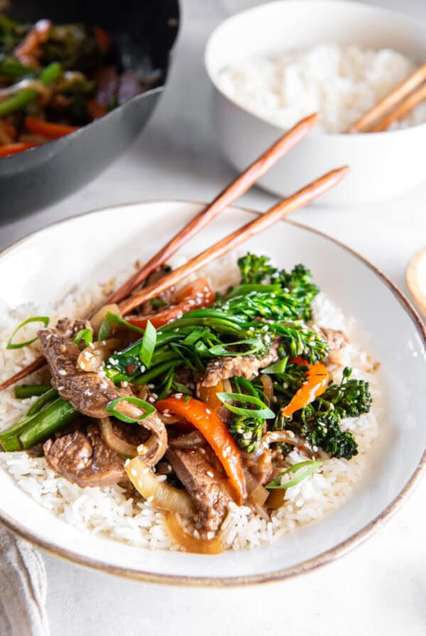 Close-up of beef and broccolini stir fry served over white rice in a shallow bowl, garnished with green onions and sesame seeds, with chopsticks resting on top.