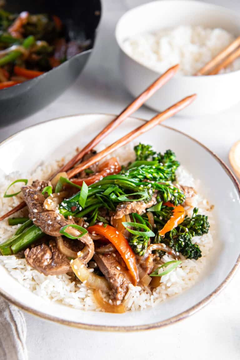 Close-up of beef and broccolini stir fry served over white rice in a shallow bowl, garnished with green onions and sesame seeds, with chopsticks resting on top.
