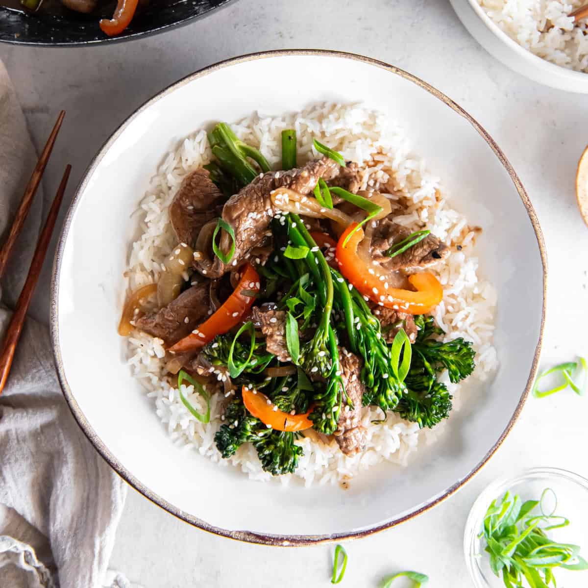 Overhead view of beef and broccolini stir fry served over white rice in a rustic bowl, topped with sliced green onions and sesame seeds.