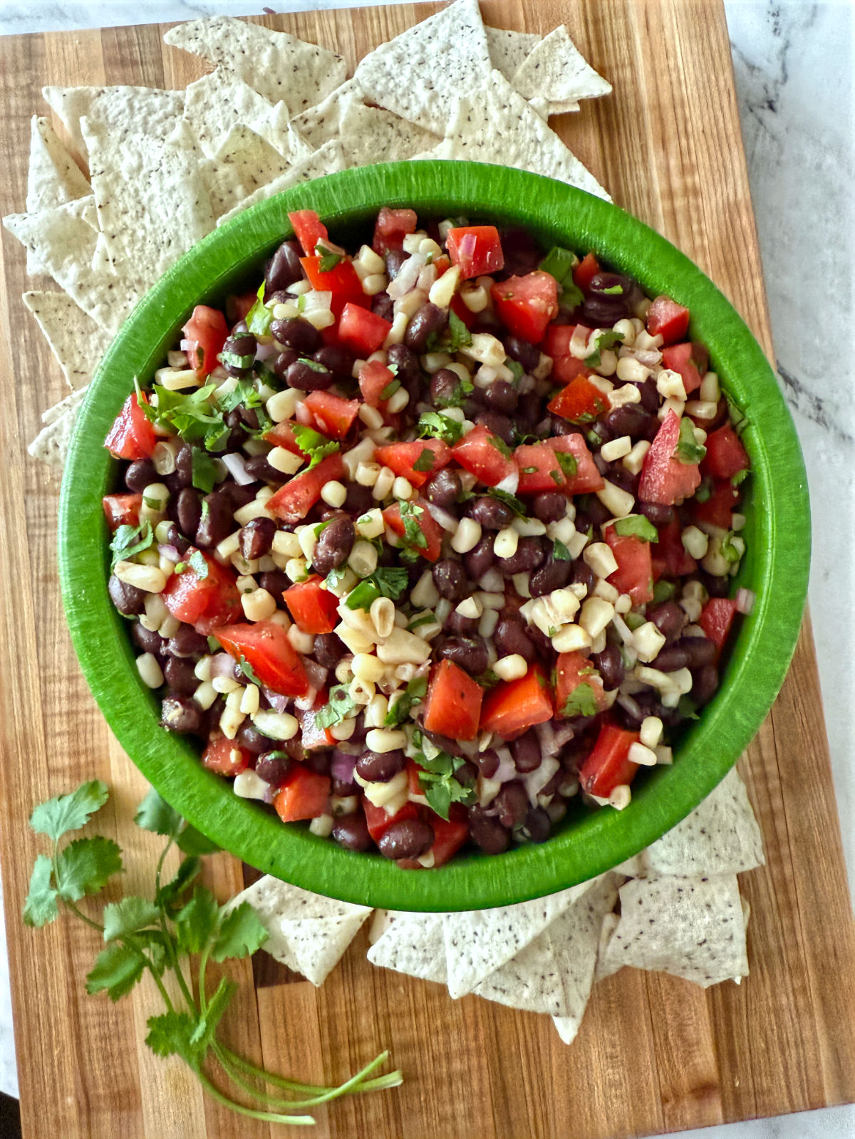 Overhead view of a green bowl filled with black bean, corn, tomato, and cilantro dip, surrounded by tortilla chips on a wooden board.