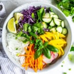 Square overhead image of a fresh spring roll bowl filled with rice noodles, shredded carrots, red cabbage, cucumber, mango, radish, fresh basil, cilantro, and lime wedges.