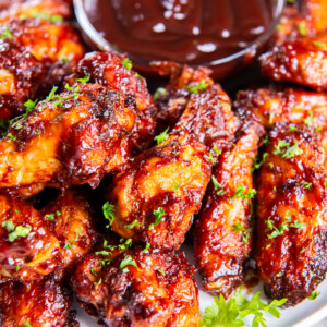 Side angle of crispy, saucy air fryer bbq chicken wings on a white plate with a bowl of barbecue sauce, garnished with fresh parsley.
