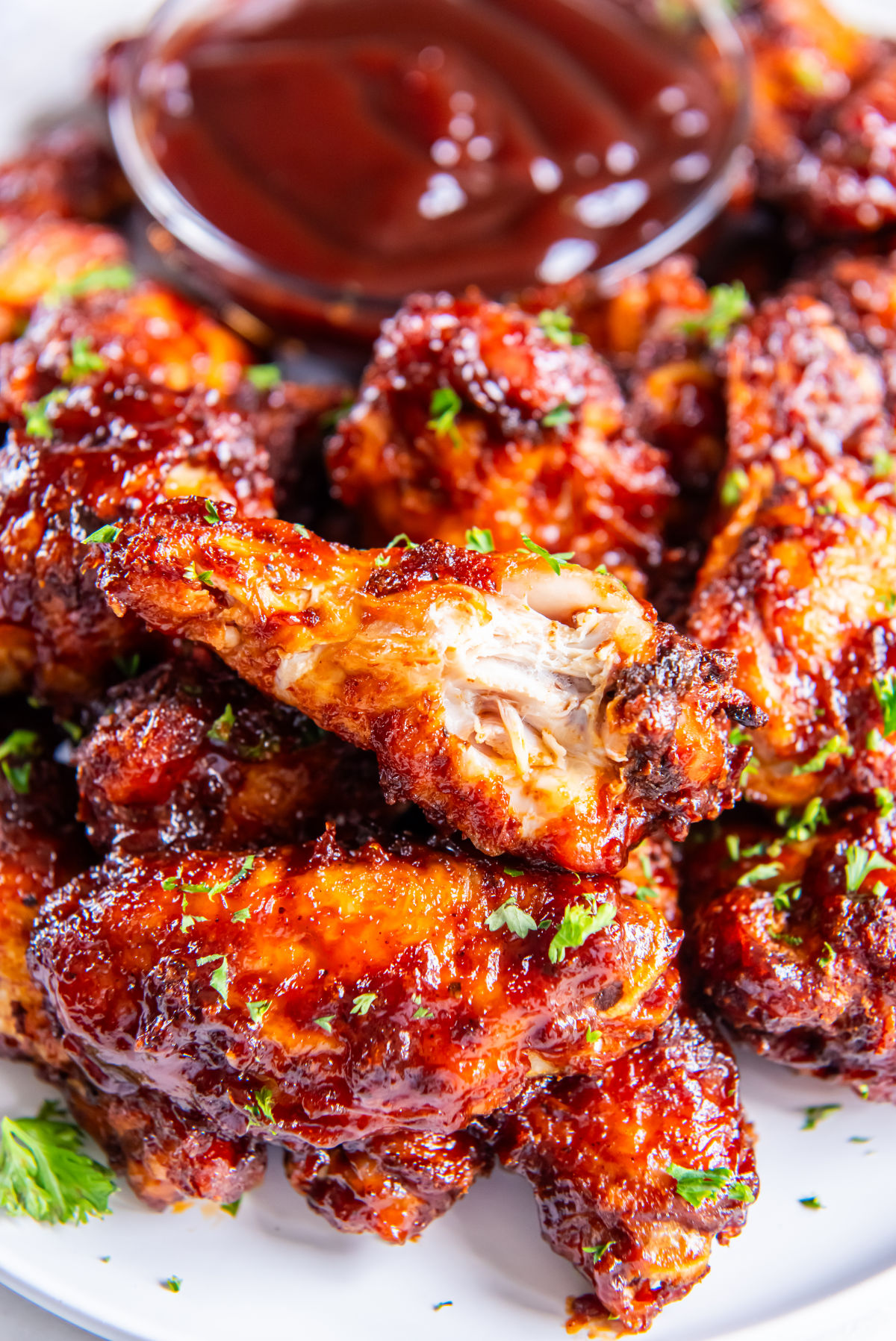 Close-up of saucy, tender air fryer bbq chicken wings with a bite taken out, revealing juicy meat inside, with BBQ sauce in the background.
