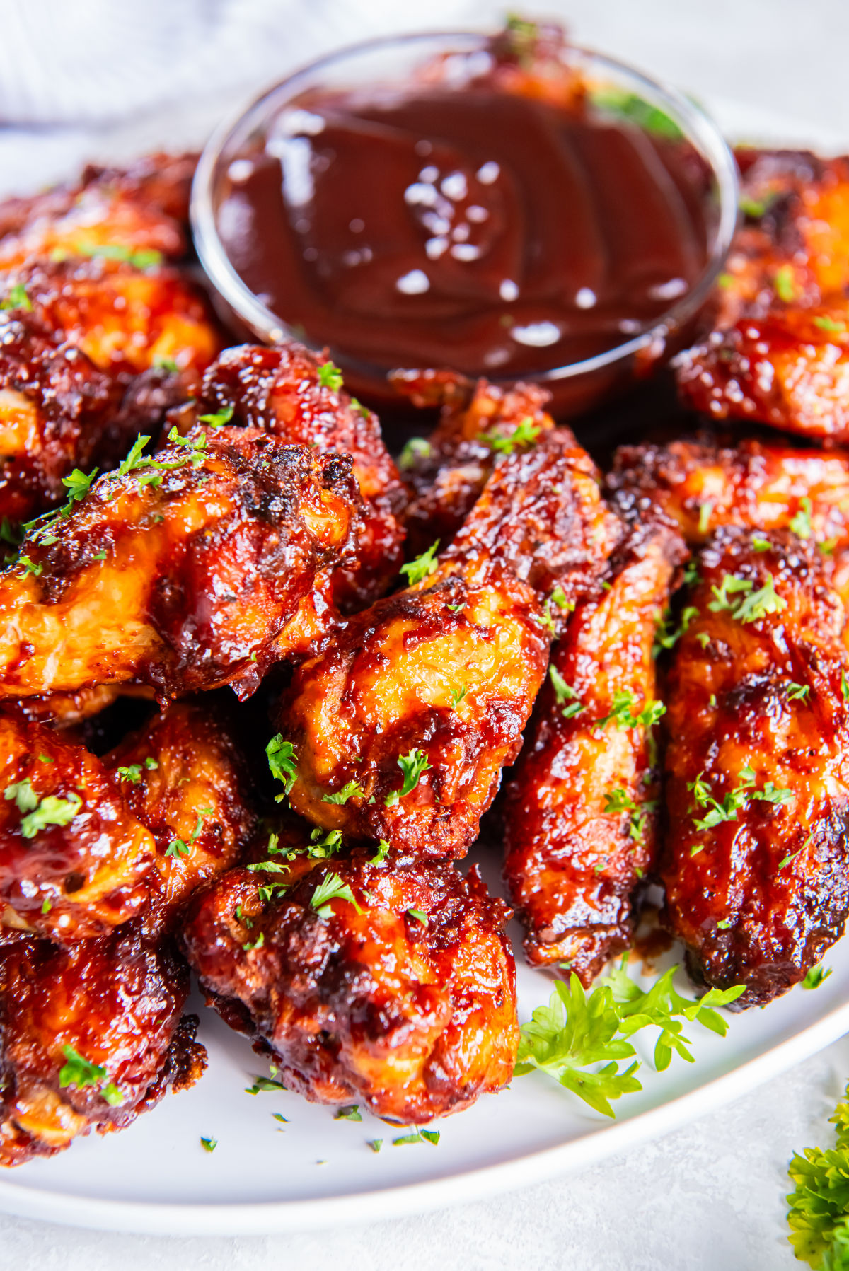 Side angle of crispy, saucy wings on a white plate with a bowl of barbecue sauce, garnished with fresh parsley.