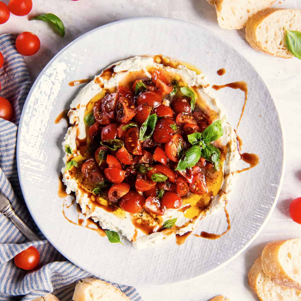 Overhead view of bruschetta dip topped with cherry tomatoes, basil, and balsamic glaze, surrounded by slices of bread.