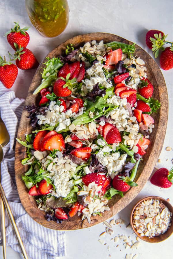 Overhead shot of salad on a wooden serving bowl surrounded by fresh strawberries, goat cheese, vinaigrette, and almond slices.