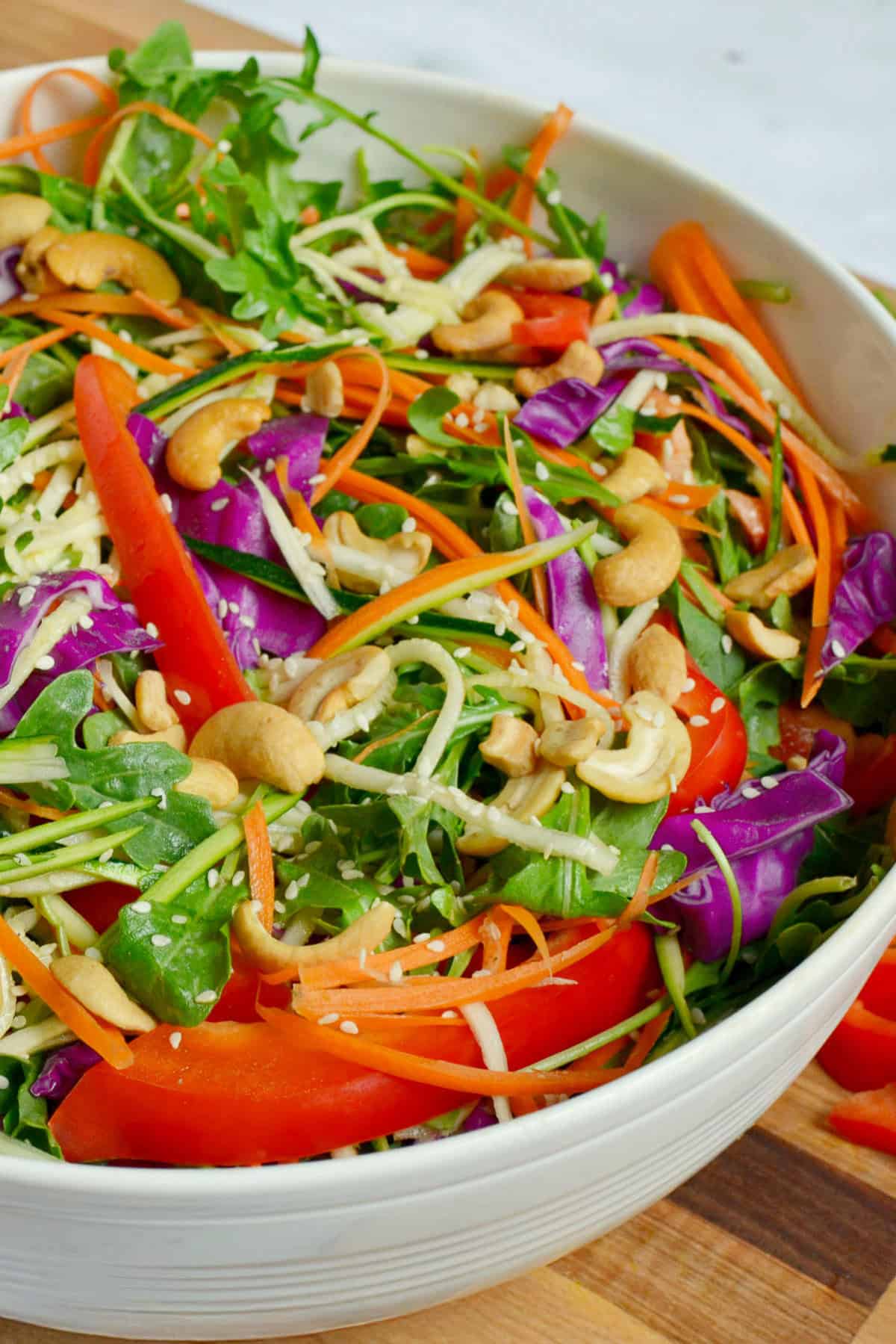 Close-up of veggie salad with carrots, red cabbage, zucchini, arugula, bell peppers, and cashews in a white bowl.