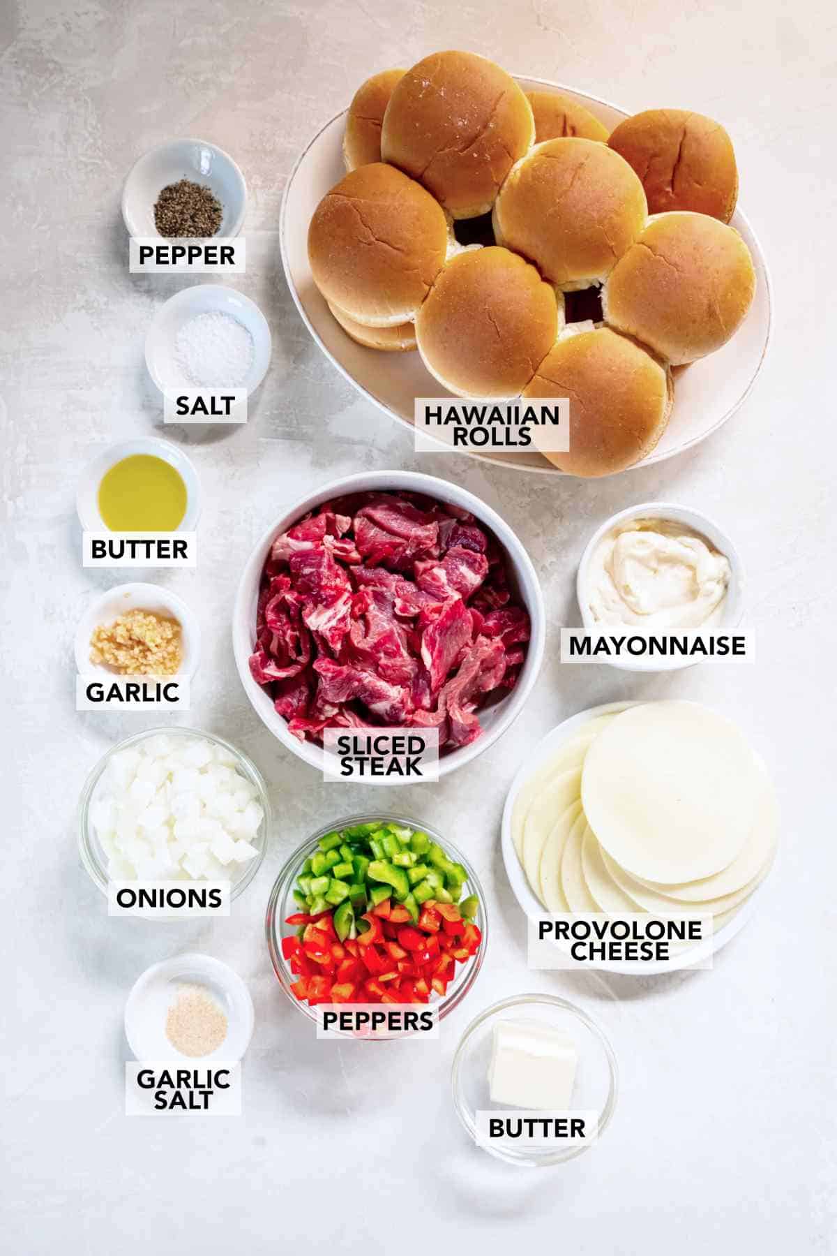 Ingredients arranged on a counter including steak, cheese, peppers, onions, and Hawaiian rolls.