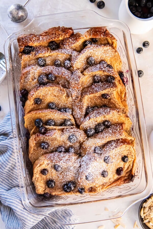 Blueberry brioche French toast bake in a glass baking dish with powdered sugar and fresh blueberries