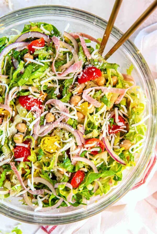 Fully tossed La Scala chopped salad with iceberg and romaine lettuce, salami, chickpeas, mozzarella, tomatoes, red onion, and pepperoncinis in a glass bowl.