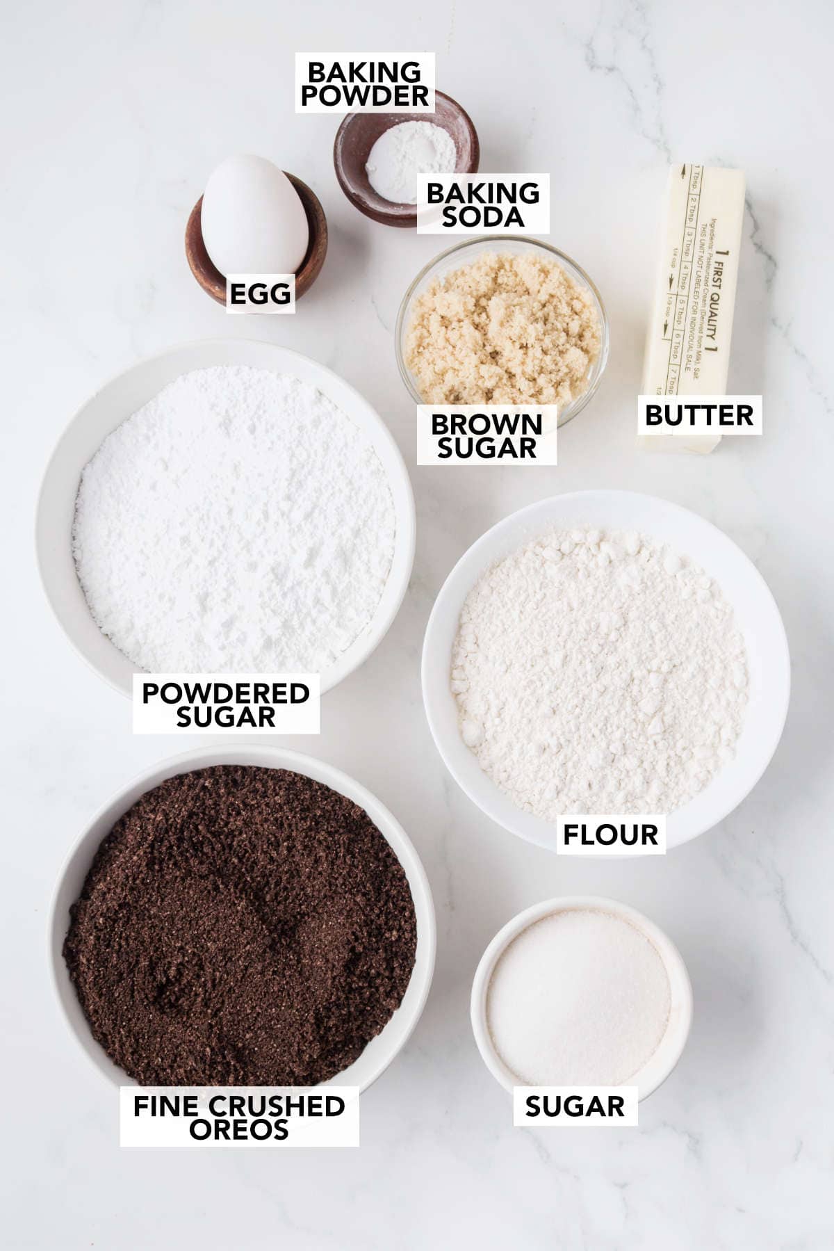 Flat lay of ingredients for Oreo Milkshake Cookies including flour, sugar, brown sugar, egg, butter, baking soda, baking powder, powdered sugar, and finely crushed Oreos.