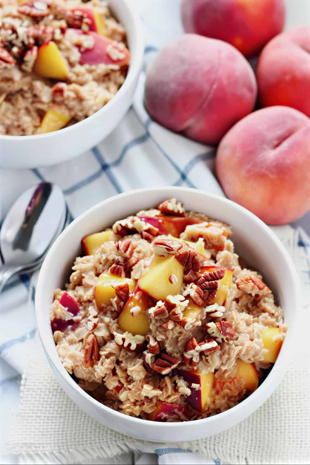 Two bowls of peach cobbler oatmeal topped with fresh peaches and pecans with whole peaches in the background.