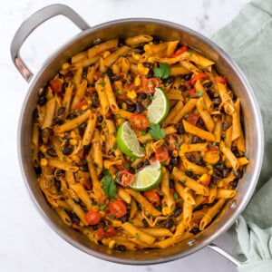 One pot chicken enchilada pasta in a skillet with black beans, corn, tomatoes and lime wedges