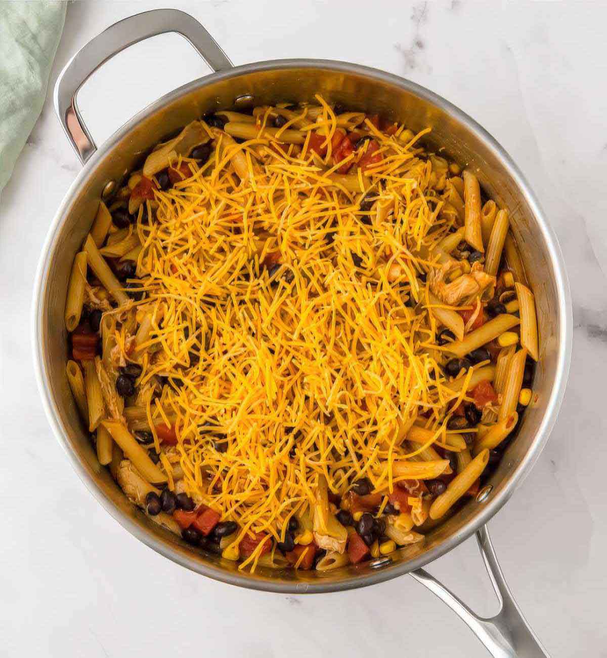 Shredded cheddar cheese sprinkled over cooked chicken enchilada pasta in a skillet