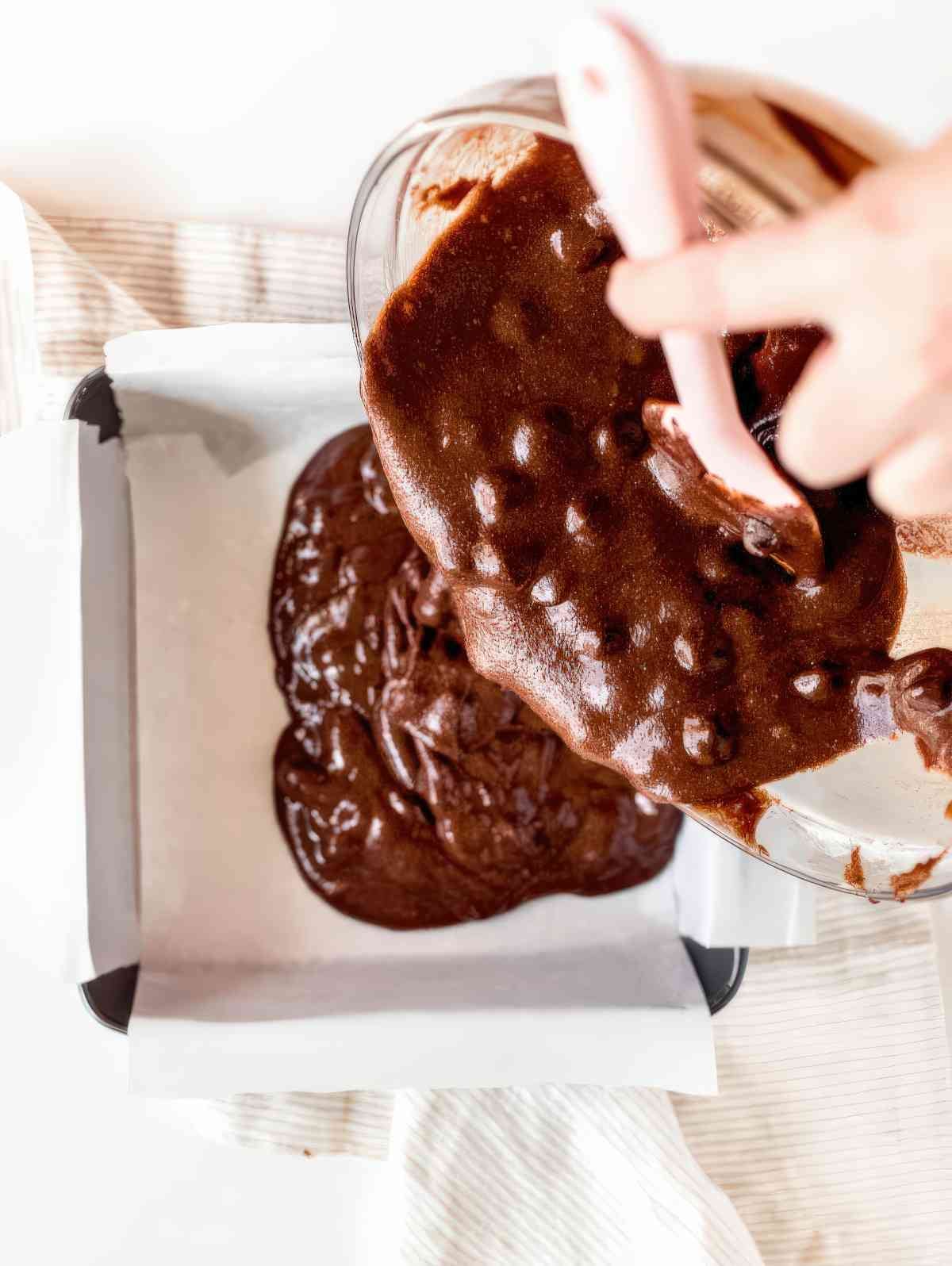 Pouring thick chocolate brownie batter into a lined baking pan