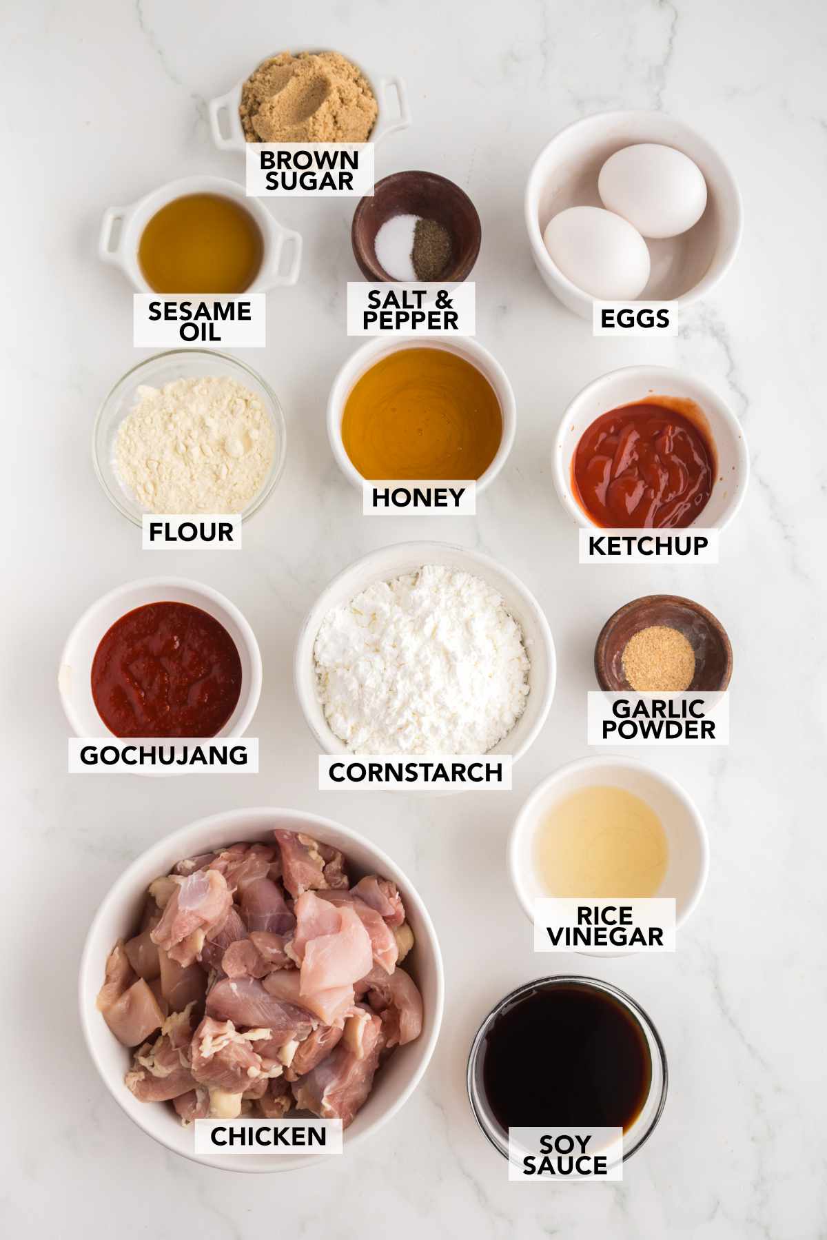 Ingredients for Crispy Gochujang Chicken including chicken thighs, gochujang, soy sauce, cornstarch, honey, sesame oil, eggs, and seasonings arranged on a white surface.