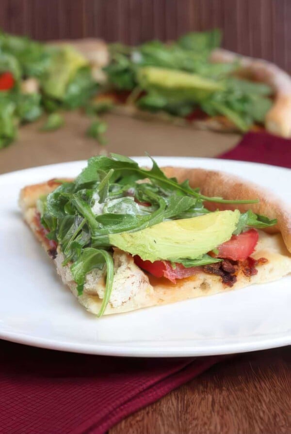 Slice of California BLT pizza topped with chicken, bacon, tomato, avocado and arugula on a white plate.