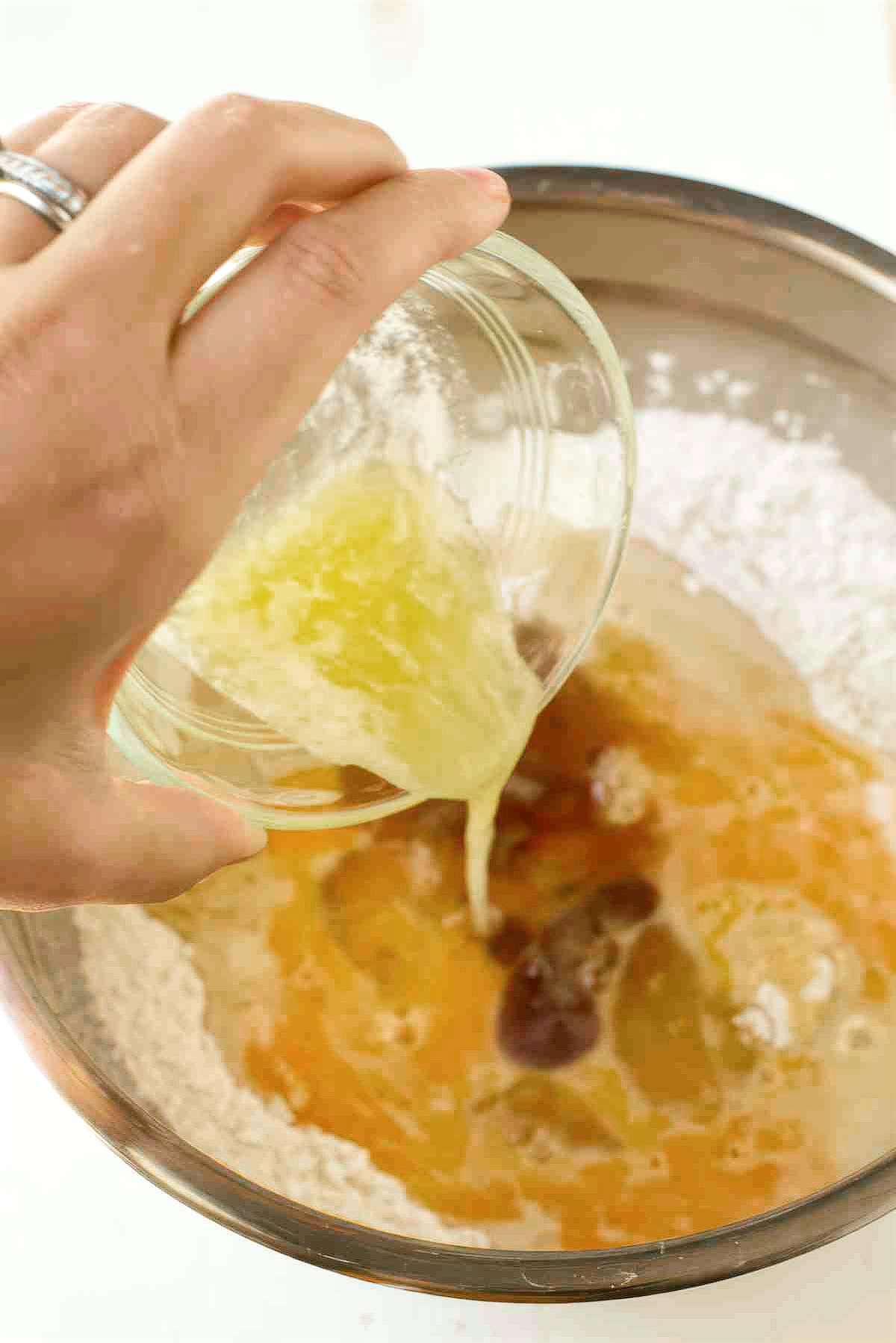 Pouring melted butter into a bowl with flour, eggs, apple cider, and other ingredients for baked apple cider donuts.