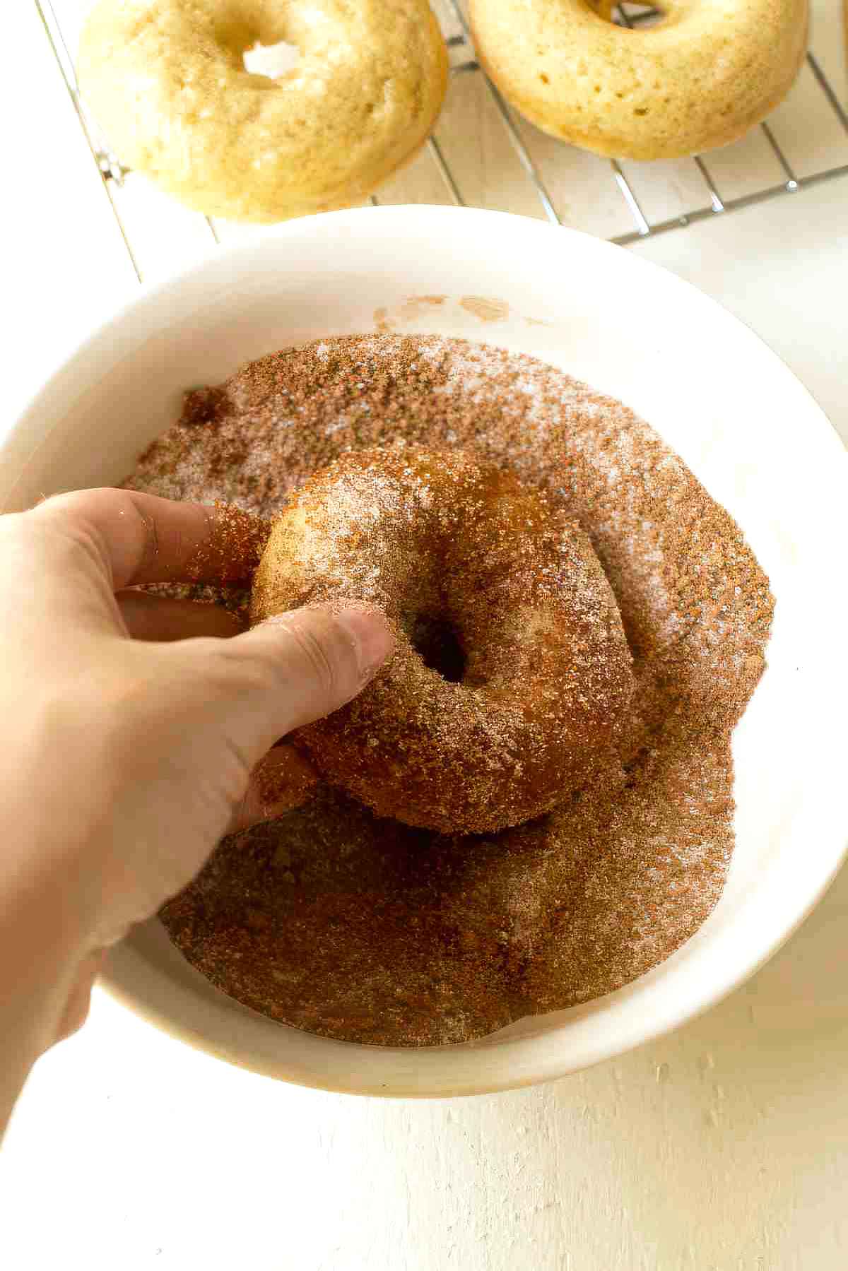 Hand coating a baked apple cider donut in cinnamon sugar with additional donuts cooling on a wire rack in the background.