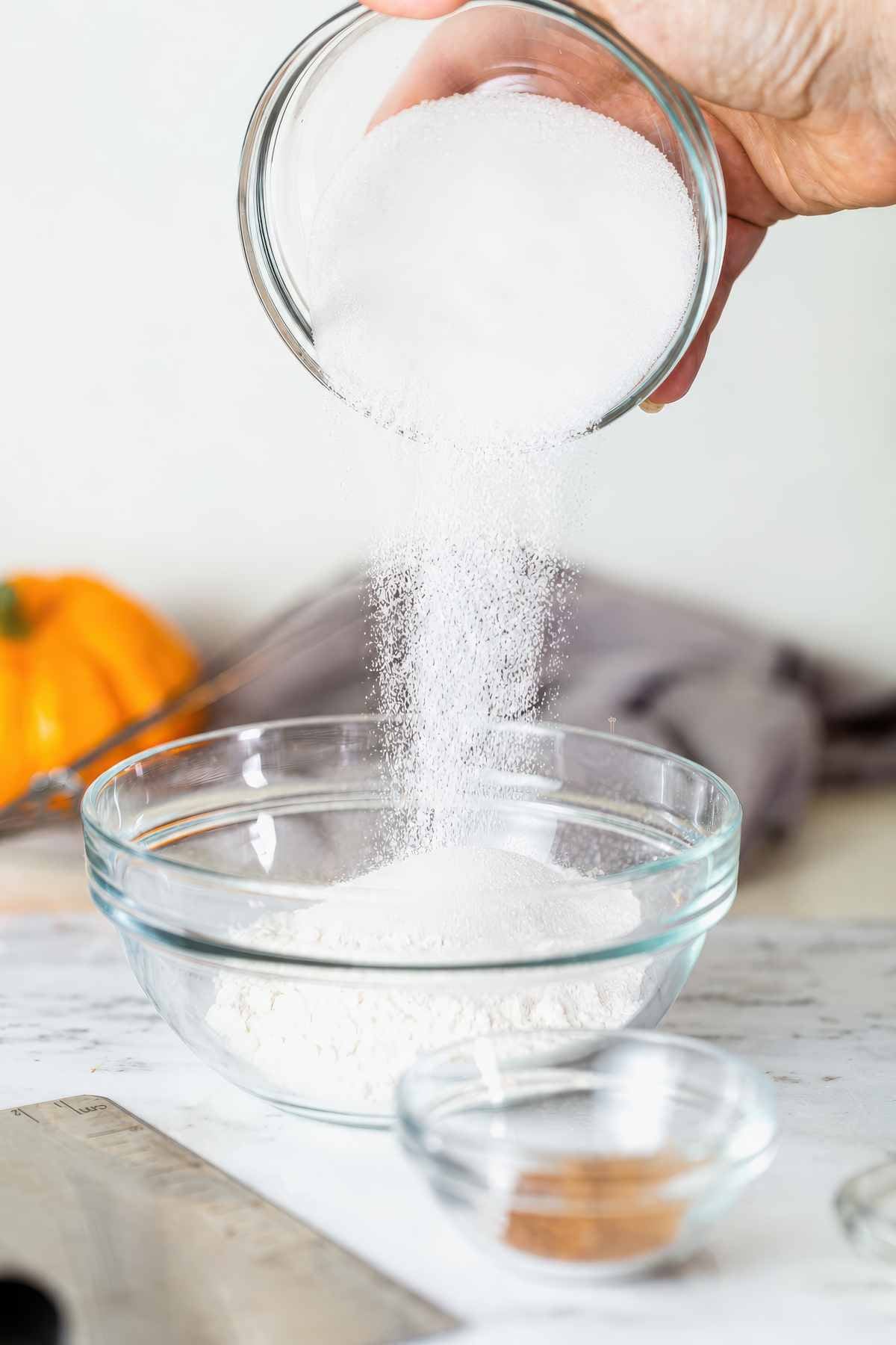 Adding granulated sugar to the dry ingredients for pumpkin muffin batter.