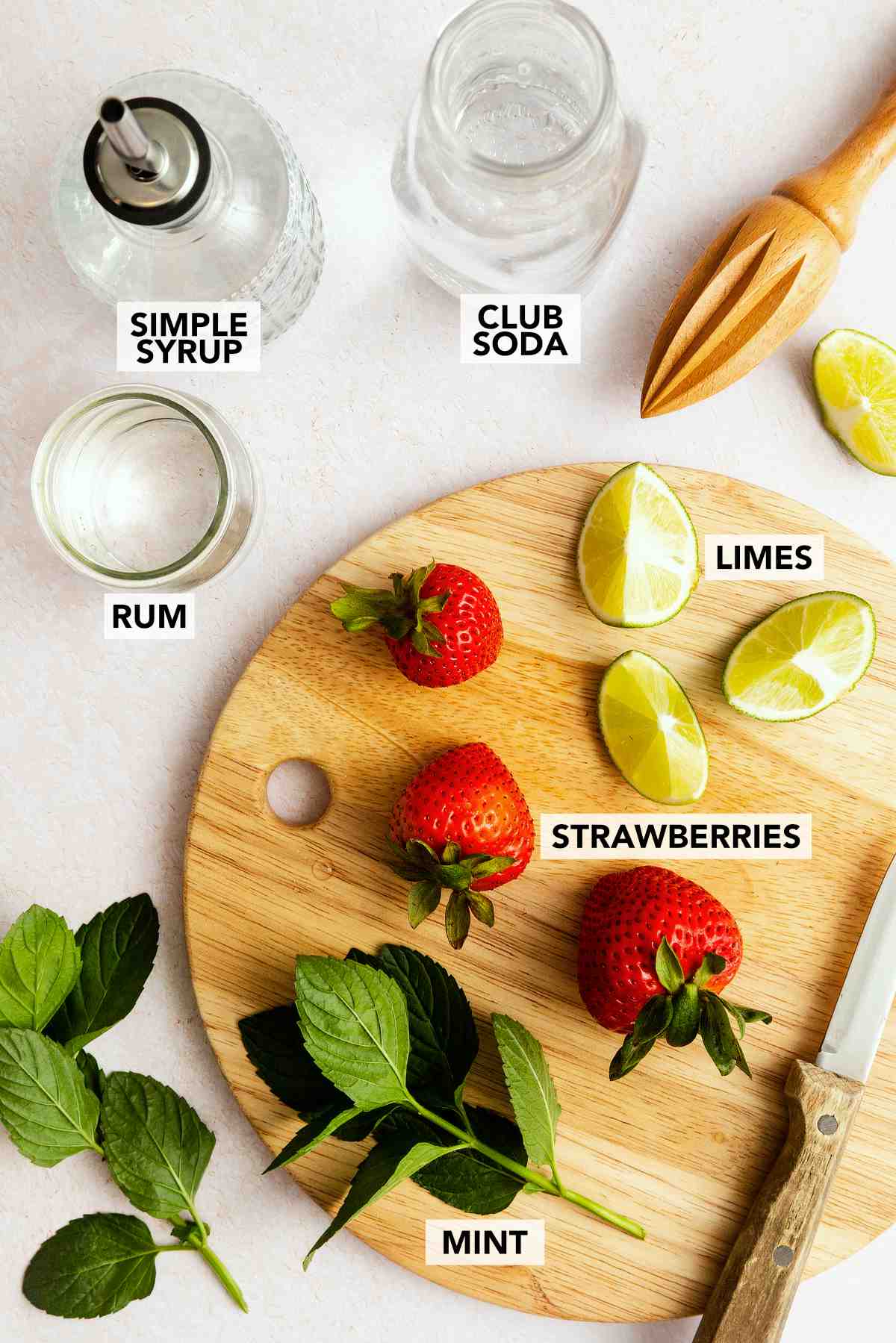 Ingredients for a strawberry mojito including fresh strawberries, mint leaves, lime wedges, rum, simple syrup, and club soda.