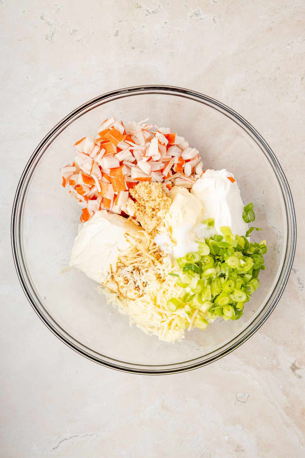 Overhead view of crab rangoon dip ingredients in a glass bowl, including chopped imitation crab, cream cheese, mozzarella, sour cream, mayonnaise, garlic, and green onions.