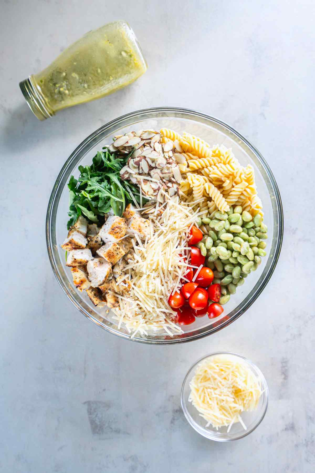 Rotini, chicken, arugula, edamame, tomatoes, almonds, and Parmesan arranged in a bowl before mixing.