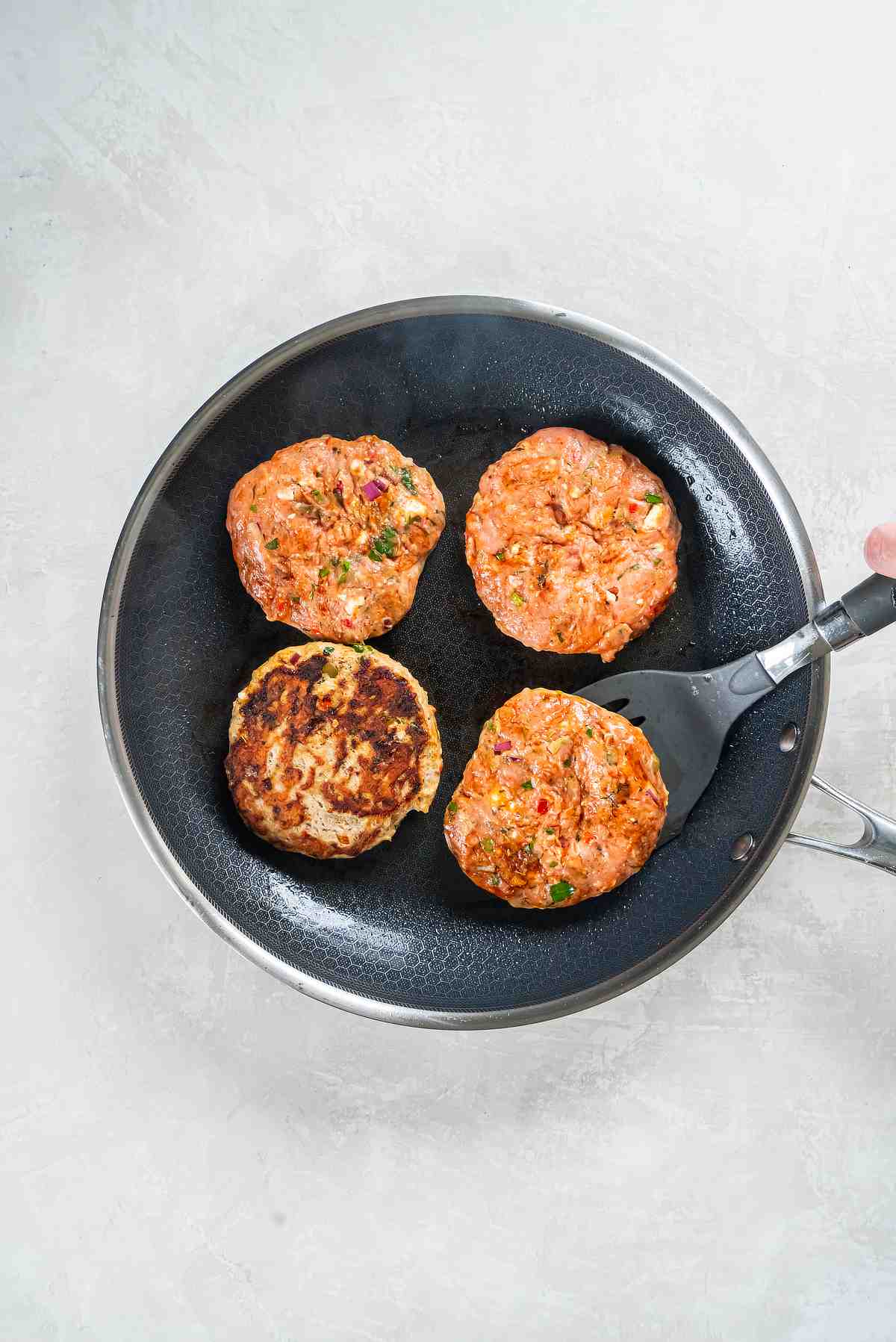 Greek turkey burger patties cooking in a skillet, with one patty flipped to show a browned crust.