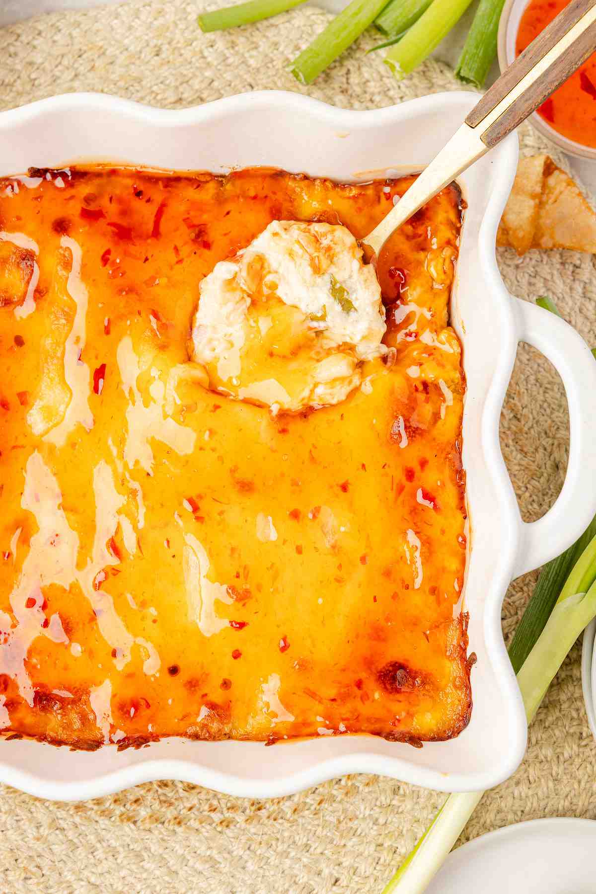 Baked crab rangoon dip being scooped with a spoon, showing melted mozzarella and a small bowl of sweet chili sauce alongside green onions.