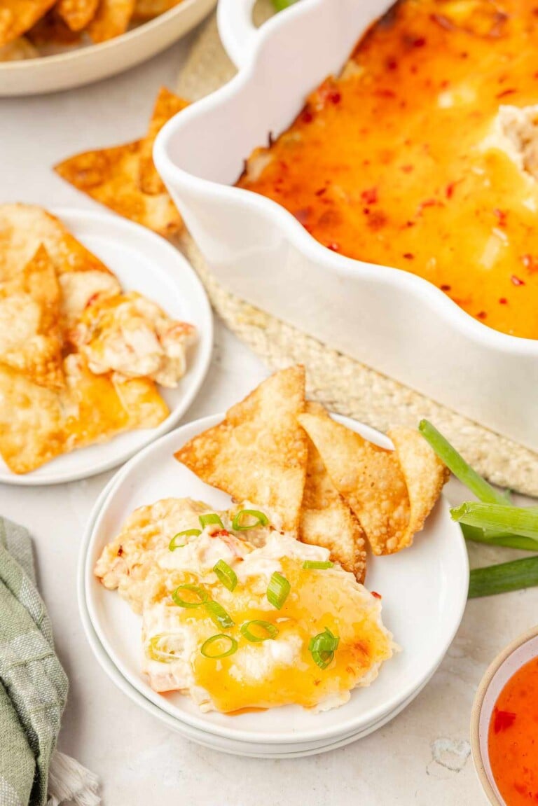 Baked crab rangoon dip in a scalloped white dish, topped with sweet chili sauce and green onions, served on small plates with crispy wonton chips.