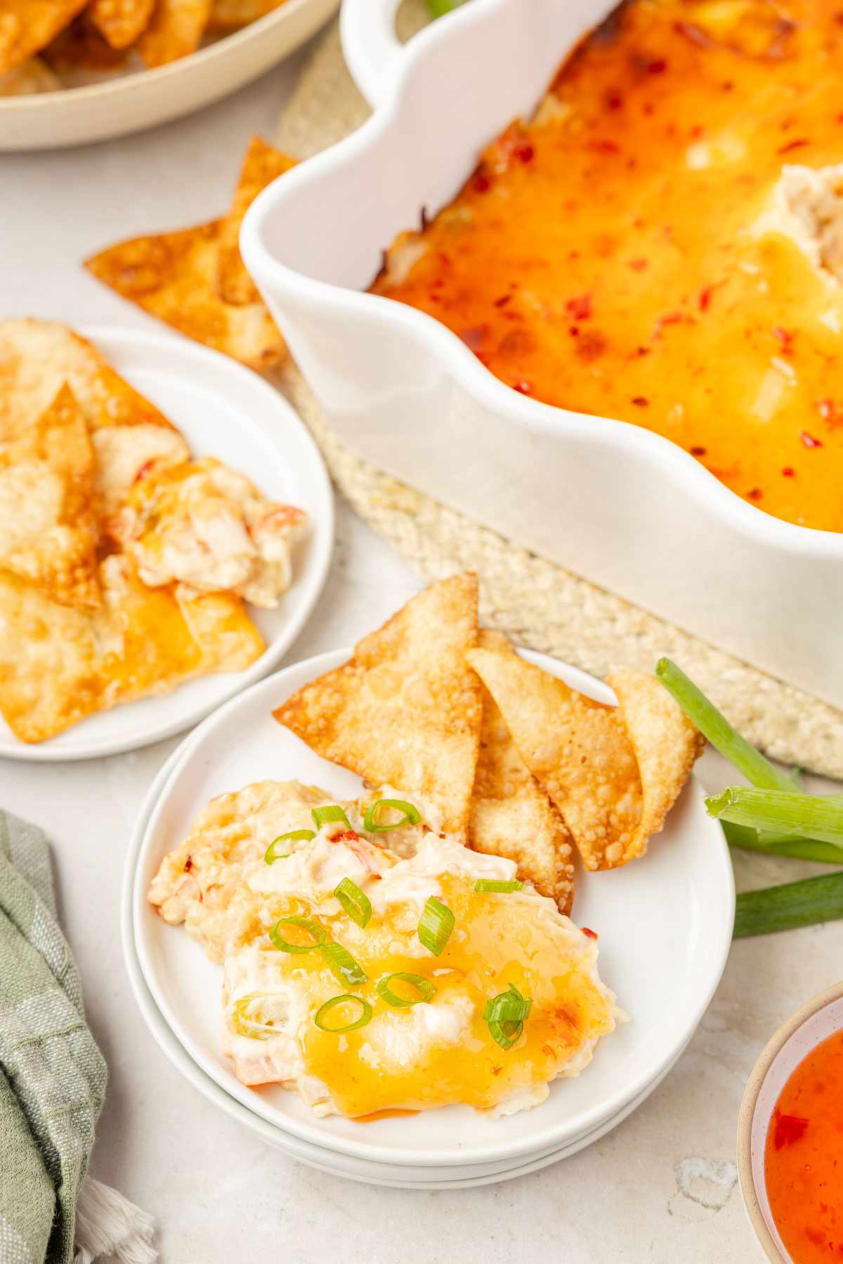 Baked crab rangoon dip in a scalloped white dish, topped with sweet chili sauce and green onions, served on small plates with crispy wonton chips.