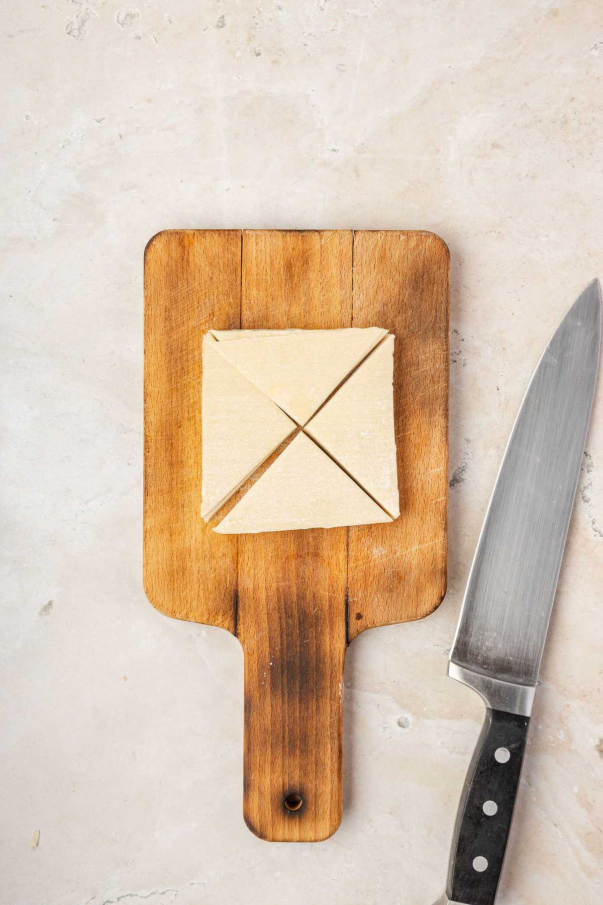 A wonton wrapper cut into four triangles on a small cutting board next to a knife, ready to fry into wonton chips.