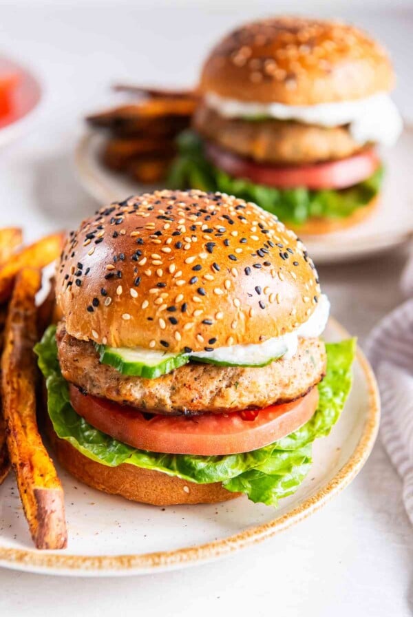 Greek turkey burger on a sesame bun with lettuce, tomato, cucumber, and tzatziki, with another burger in the background.
