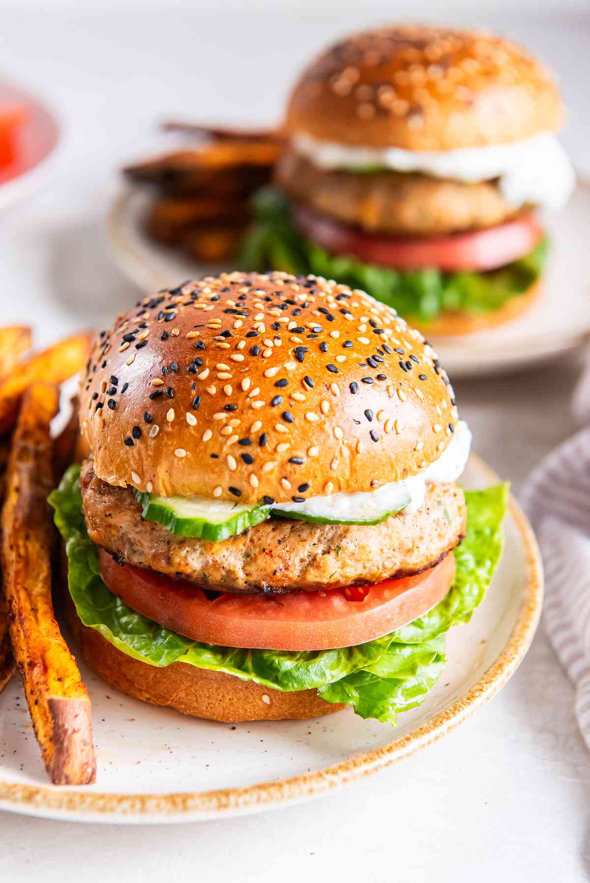 Greek turkey burger on a sesame bun with lettuce, tomato, cucumber, and tzatziki, with another burger in the background.