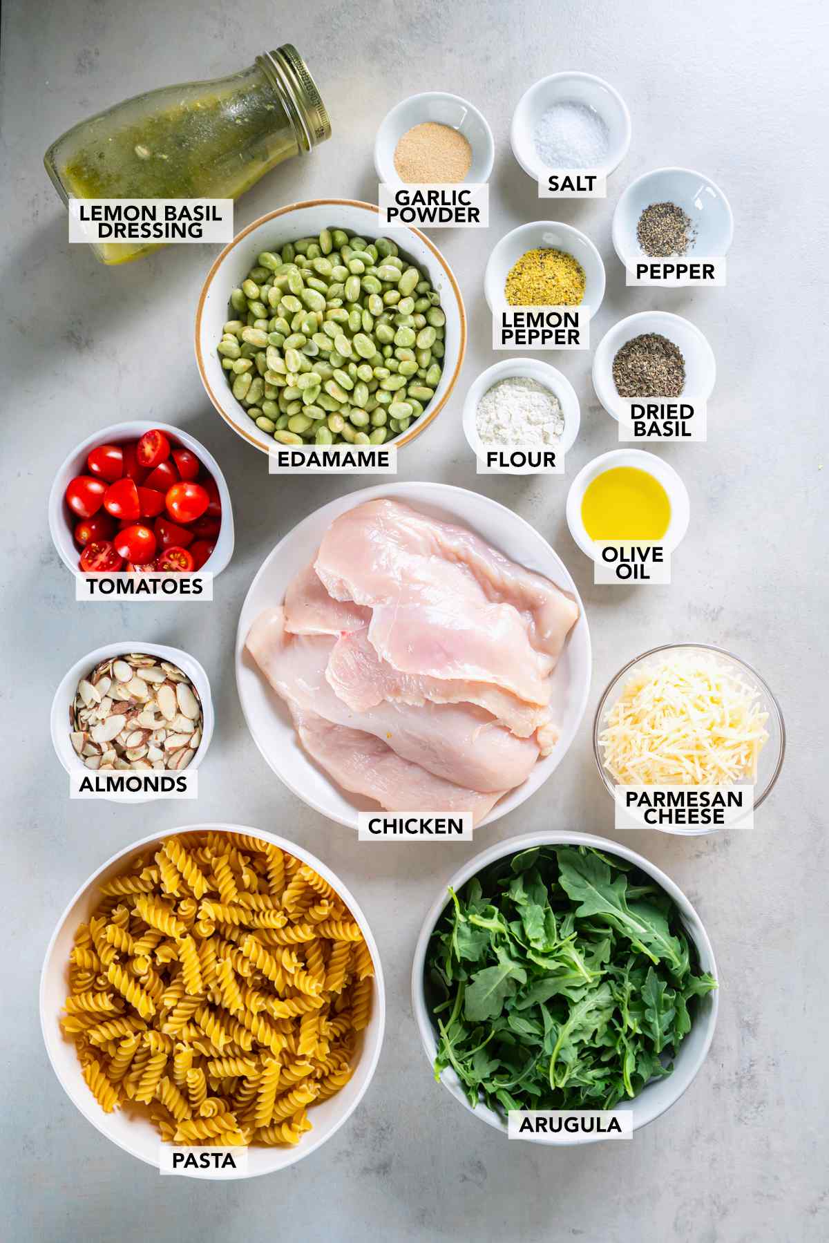 Ingredients for lemon basil chicken pasta salad, including chicken, rotini, arugula, edamame, tomatoes, almonds, Parmesan, and lemon basil dressing.