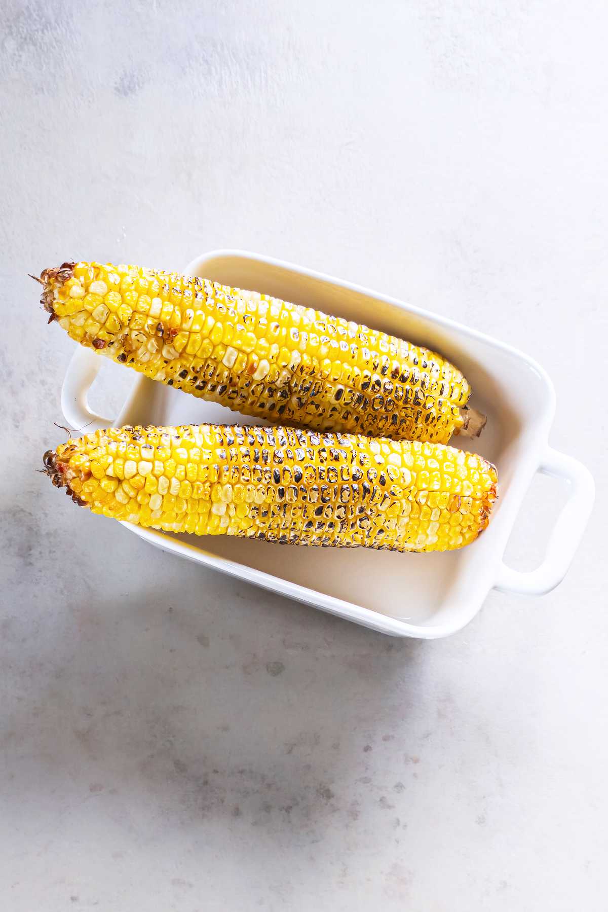 Two grilled ears of corn with browned char marks resting in a white baking dish.