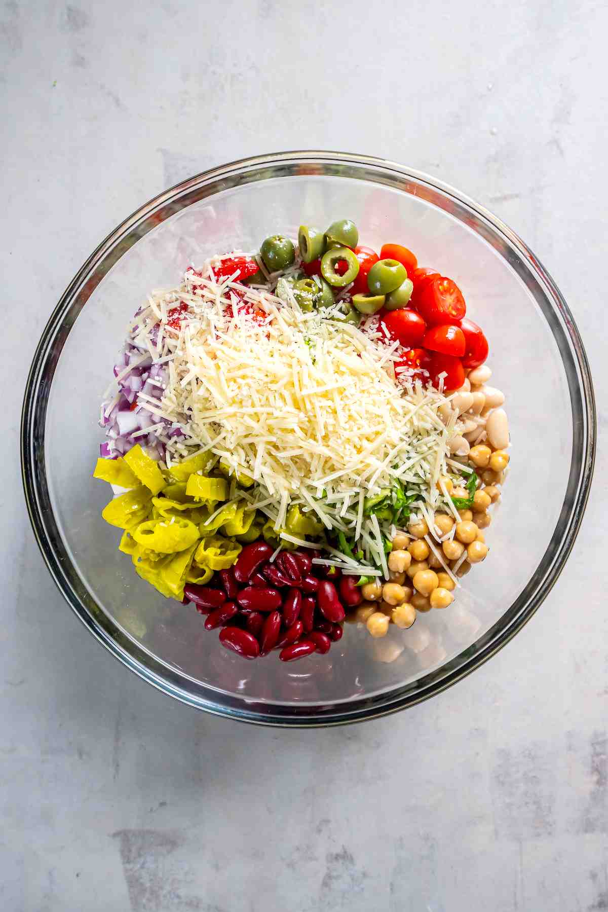 Three beans, tomatoes, red onion, olives, roasted red peppers, pepperoncini, herbs, and Parmesan in a glass mixing bowl.