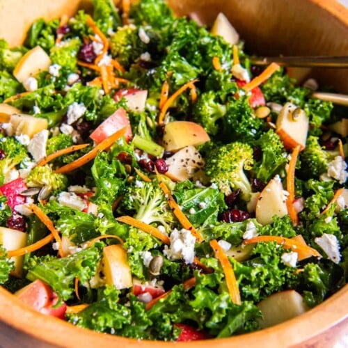 Healthy kale apple salad with broccoli, carrots, feta, dried cranberries, and pepitas in a wooden bowl.