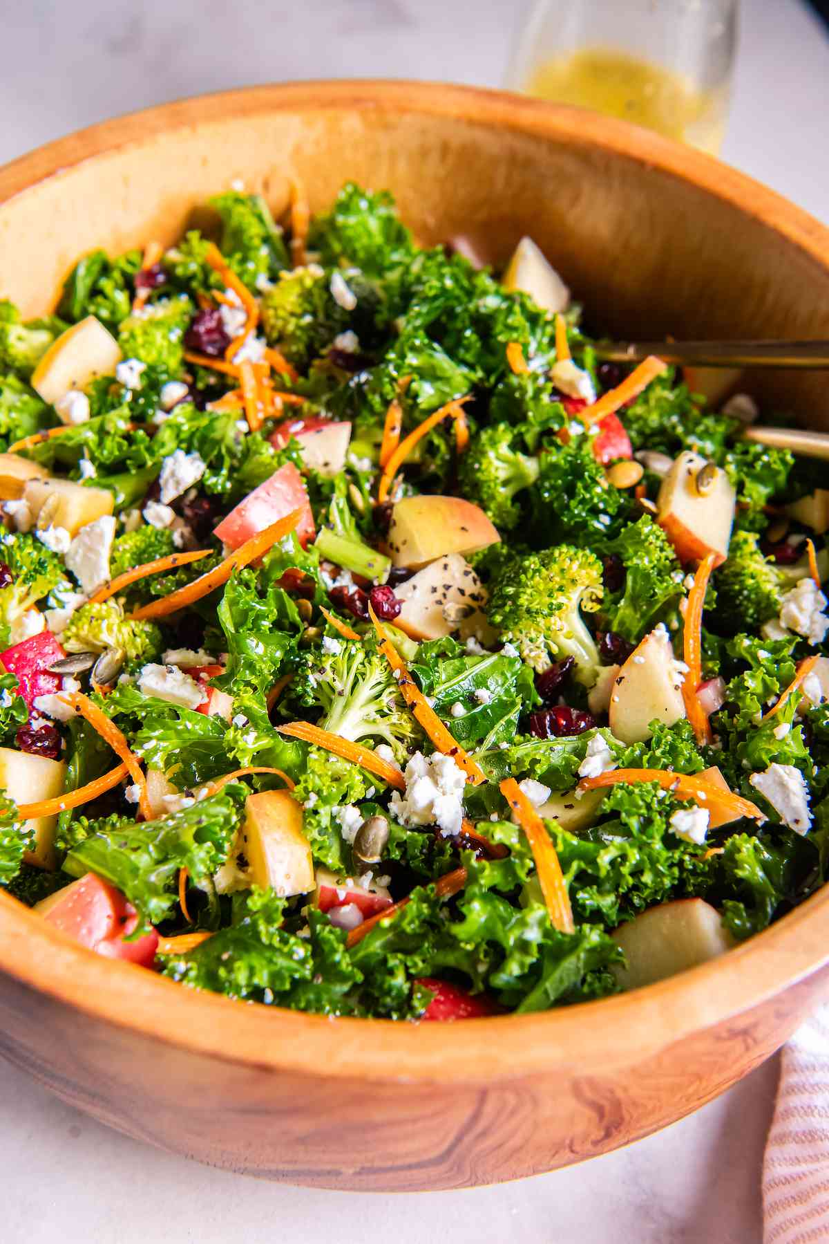 Healthy kale apple salad with broccoli, carrots, feta, dried cranberries, and pepitas in a wooden bowl.