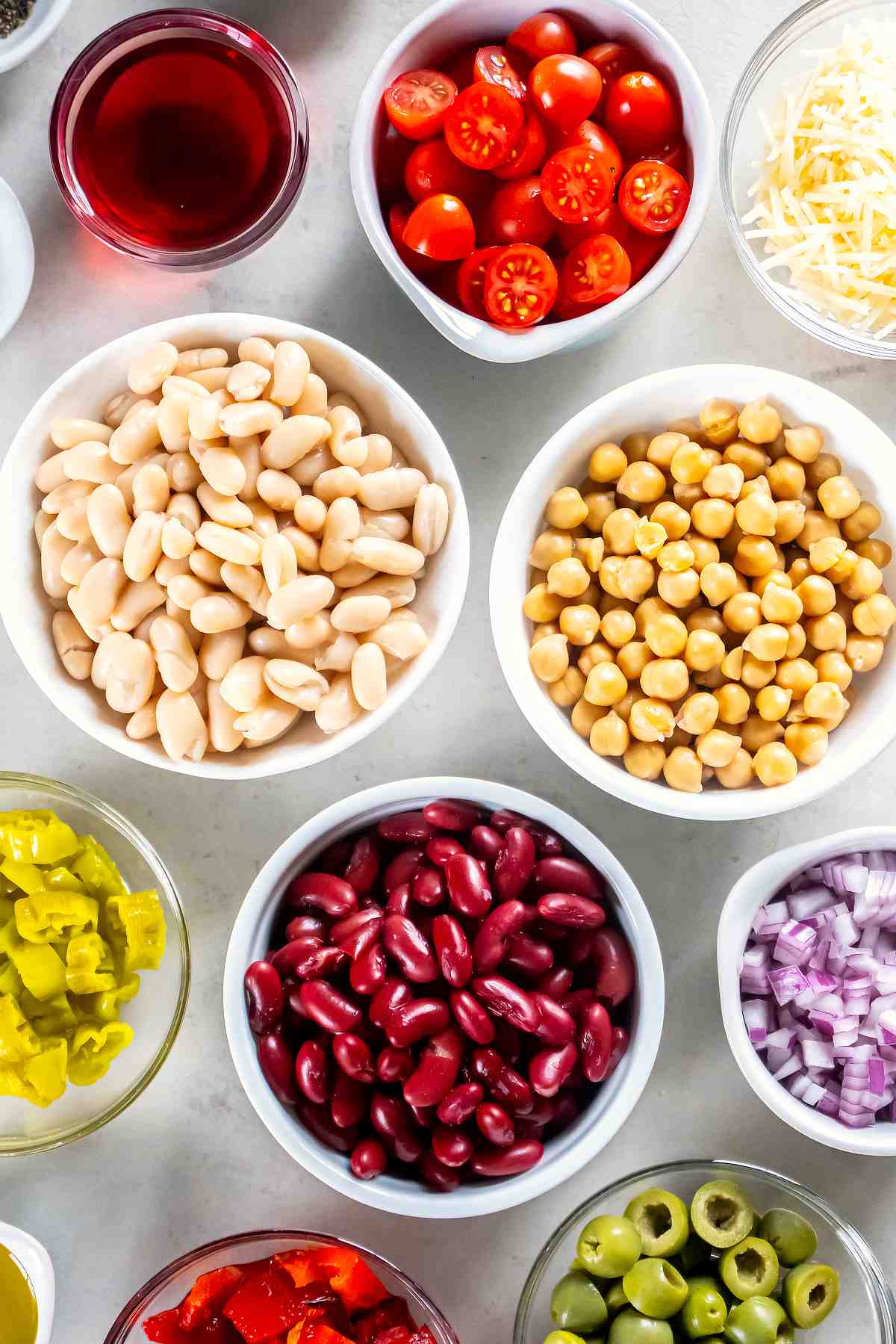 Chickpeas, cannellini beans, kidney beans, tomatoes, red onion, olives, pepperoncini, roasted red peppers, Parmesan, and red wine vinegar arranged in bowls.