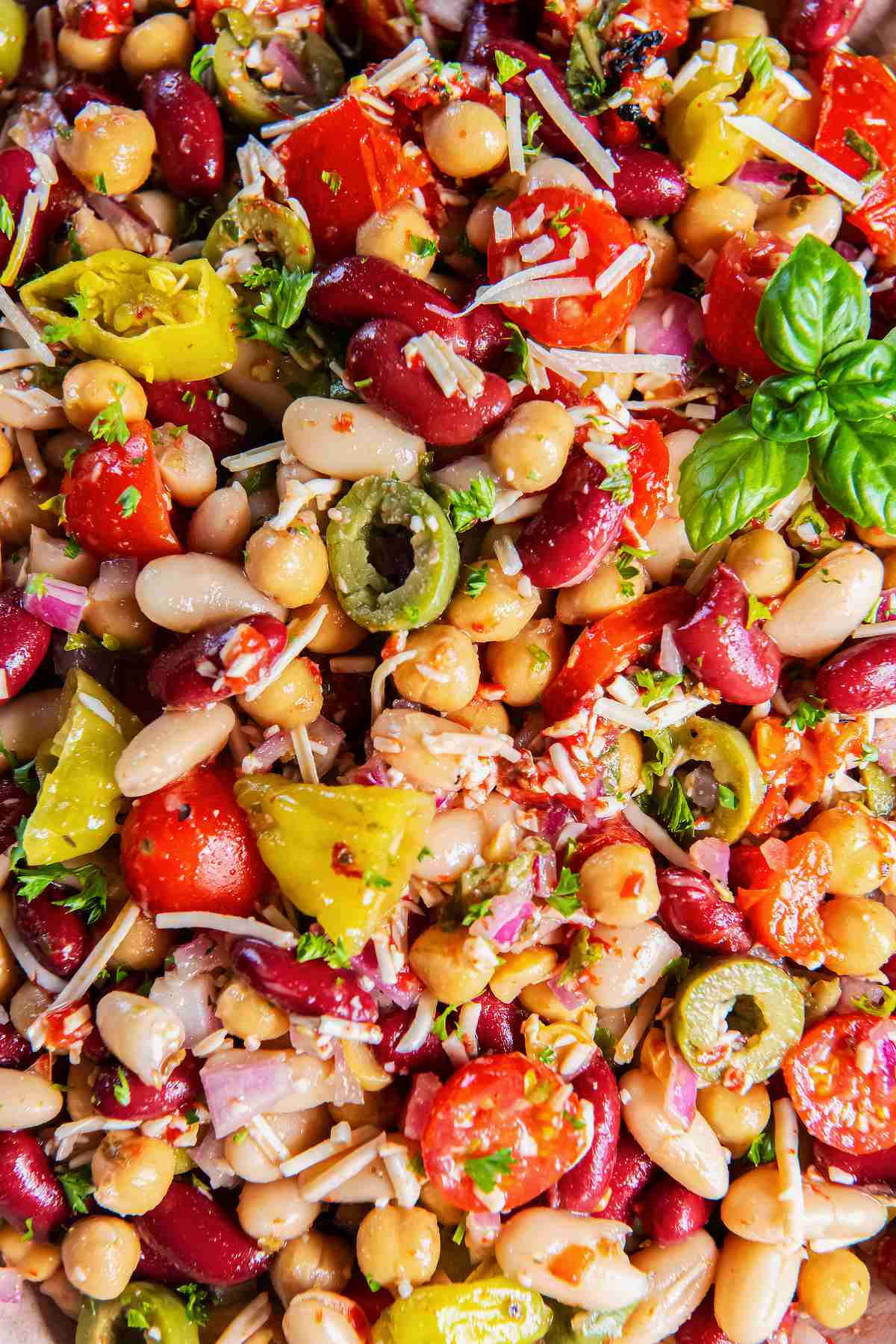 Close-up of Italian bean salad with three kinds of beans, tomatoes, roasted red peppers, olives, pepperoncini, Parmesan, and basil.