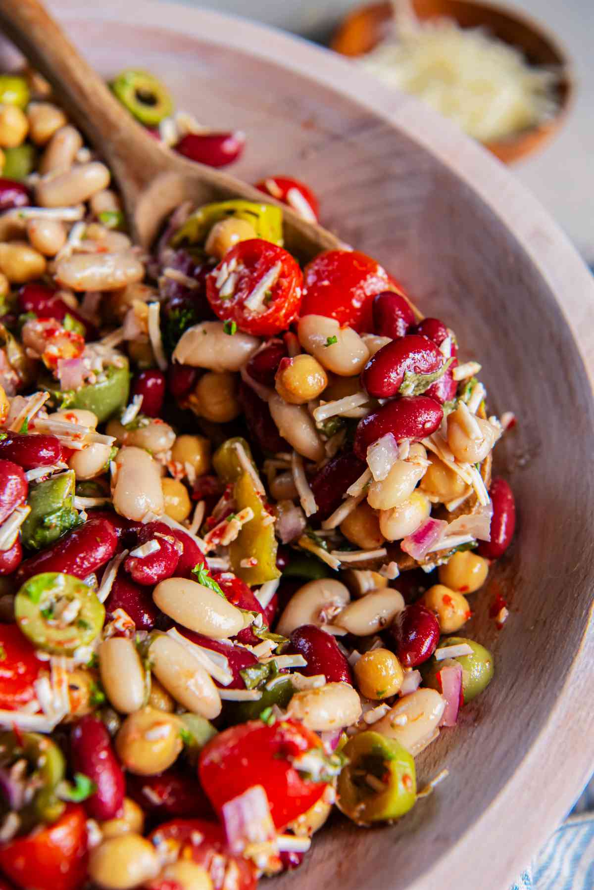 Wooden spoon lifting Italian three bean salad with creamy beans, cherry tomatoes, olives, red onion, pepperoncini, and Parmesan.