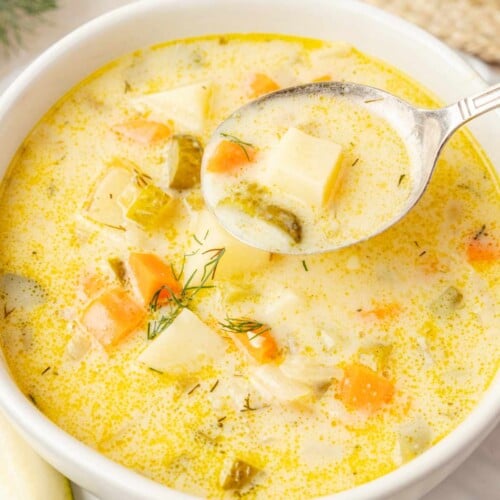 Spoon holding a potato chunk, carrot and pickle in creamy broth above a bowl of dill pickle soup.