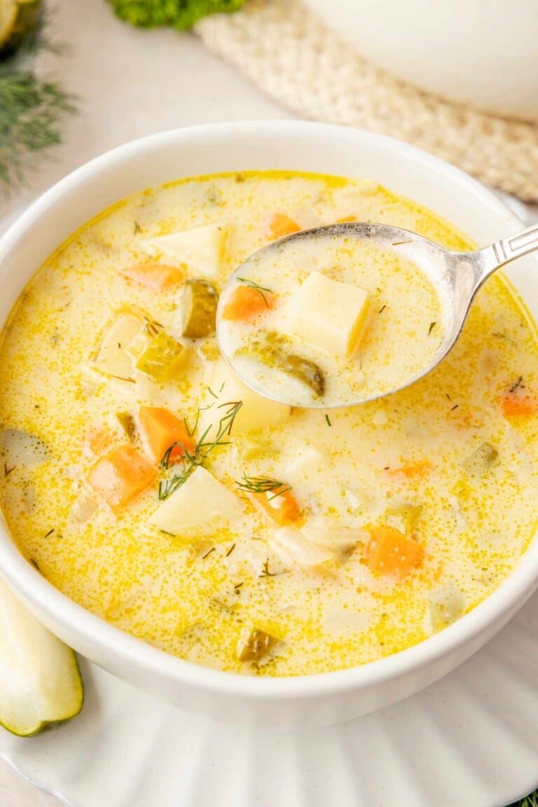 Spoon holding a potato chunk, carrot and pickle in creamy broth above a bowl of dill pickle soup.