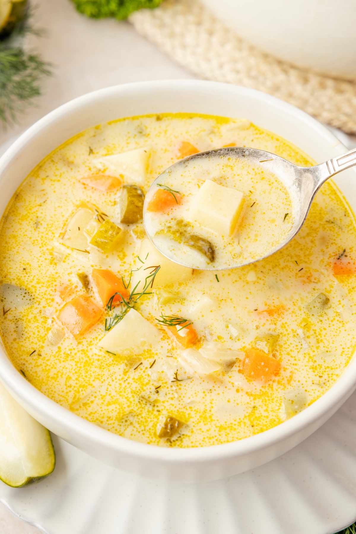 Spoon holding a potato chunk, carrot and pickle in creamy broth above a bowl of dill pickle soup.