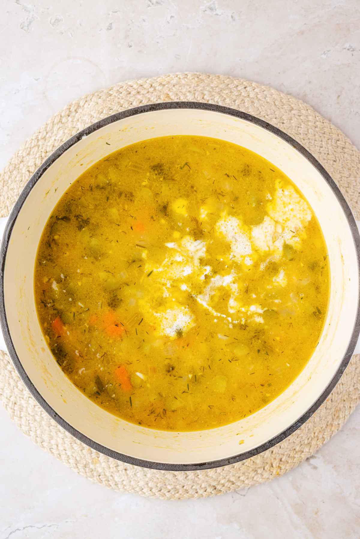 White cream pooling on the surface of dill pickle soup with potatoes, carrots and dill in a Dutch oven.